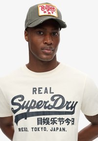 Camiseta crema con texto gráfico en azul marino "REAL SuperDry" y caracteres japoneses. Gorra oliva con parche de logo bordado. Textura de algodón suave.