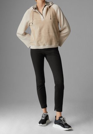 Tarnfarbener und cremefarbener Pullover-Hoodie mit einer Fronttasche, gerippten Bündchen und einem gemusterten Kragen. Kombiniert mit schwarzen, figurbetonten Hosen und schwarzen Turnschuhen.