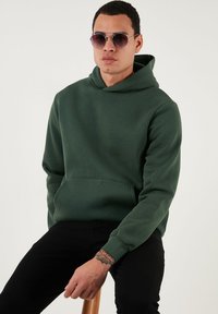 Zelená mikina s kapucňou a predným vreckom, vyrobená z mäkkého materiálu. Model má na sebe čierne nohavice a okrúhle slnečné okuliare.