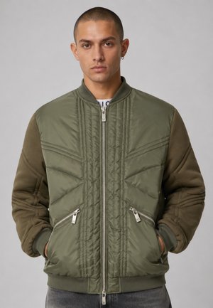 Uomo che indossa una giacca bomber imbottita verde con tasche con zip, in piedi con le mani nelle tasche su uno sfondo grigio neutro.