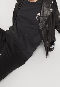 Černá kožená motorkářská bunda se stříbrnými zipy, nošená přes černou mikinu s oblíbeným kulatým logem v metalické zlaté a bílé barvě.