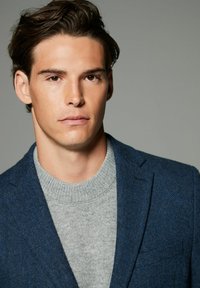 Blauer Fischgrat-Blazer über einem hellgrauen Strickpullover. Der Blazer hat einen Reverskragen und eine taillierte Passform, die durch den strukturierten Stoff hervorgehoben wird.