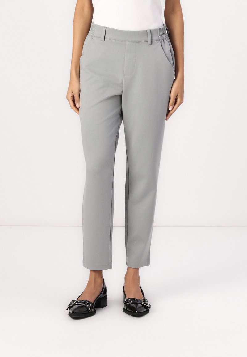 Femme portant un pantalon gris clair sur mesure, longueur cheville, avec poches latérales et des chaussures noires en cuir verni à petits talons avec boucles.