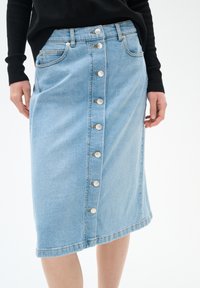Jupe en denim bleu clair, longueur genoux, avec une fermeture frontale à boutons, deux poches latérales et des coutures visibles le long de l'ourlet.