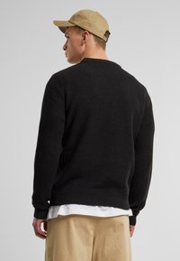 Black ribbed knit sweater with a crew neckline, long sleeves, and a relaxed fit, layered over a white shirt, paired with beige pants.
