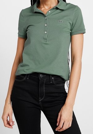 Kvinde iført en grøn kortærmet poloshirt med knapper og sorte højtaljede jeans mod en ensfarvet baggrund.