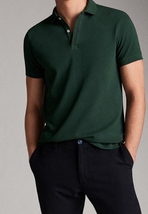 Homme portant un polo à manches courtes vert foncé et un pantalon noir, les mains dans les poches, debout devant un fond neutre.