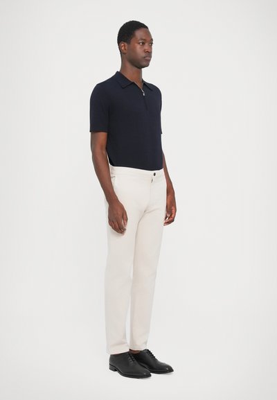 Polo shirt bleumarin cu guler cu fermoar, asociat cu pantaloni crem și pantofi negri. Textură netedă, croială ajustată și design simplu.
