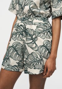 Μπεζ υφασμάτινη βερμούδα με σκούρα πράσινα περίπλοκα μοτίβα φύλλων, φορεμένη με ταιριαστή μπλούζα, δείχνοντας ελαστική μέση και μέρος του δεξιού βραχίονα.