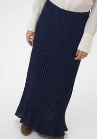 Kvinde iført en marineblå plisseret maxiskørt, hvid bluse med broderede manchetter og brune sko, stående foran en ensfarvet baggrund.