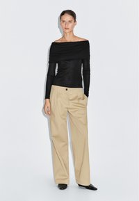 Schwarzes, schulterfreies Langarmoberteil in Kombination mit beigen, hochgeschnittenen, weiten Hosen. Schwarze, spitz zulaufende Schuhe. Glatte Stoffstrukturen sind sichtbar.