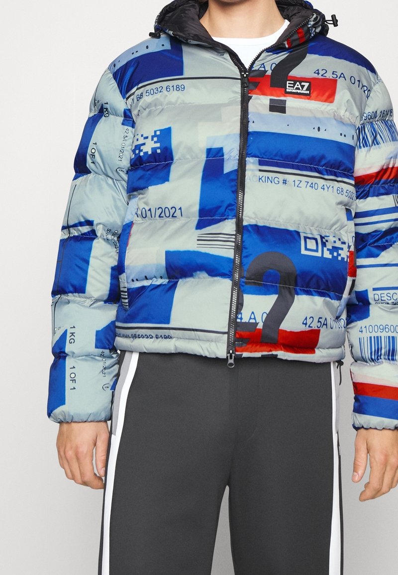 Niebieska, szara i czerwona puchowa kurtka w wzory z zamkiem z przodu i kapturem, noszona z ciemnoszarymi spodniami z białymi paskami po bokach.