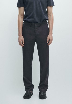 Uomo che indossa pantaloni scuri a gamba dritta, mocassini neri in pelle e una camicia scura a maniche corte, in piedi contro uno sfondo chiaro e semplice.