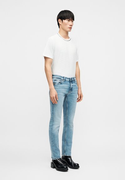 Λευκό t-shirt με στρογγυλή λαιμόκοψη, ανοιχτό μπλε τζιν skinny, και μαύρα γυαλιστερά μποτάκια με χοντρό τακούνι. Απλός σχεδιασμός, χωρίς σχέδια ή ορατά λογότυπα.