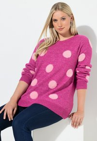 Frau mit langen blonden Haaren, die einen rosa Pullover mit hellrosa Punkten und dunkelblaue Jeans trägt, sitzt vor weißem Hintergrund.