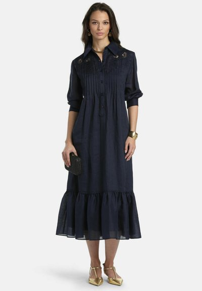 Žena v dlhej tmavomodrej šaty s výšivkou, drží čiernu kabelku, zlatý náramok a zlaté špicaté lodičky, stojí pred bielym pozadím.