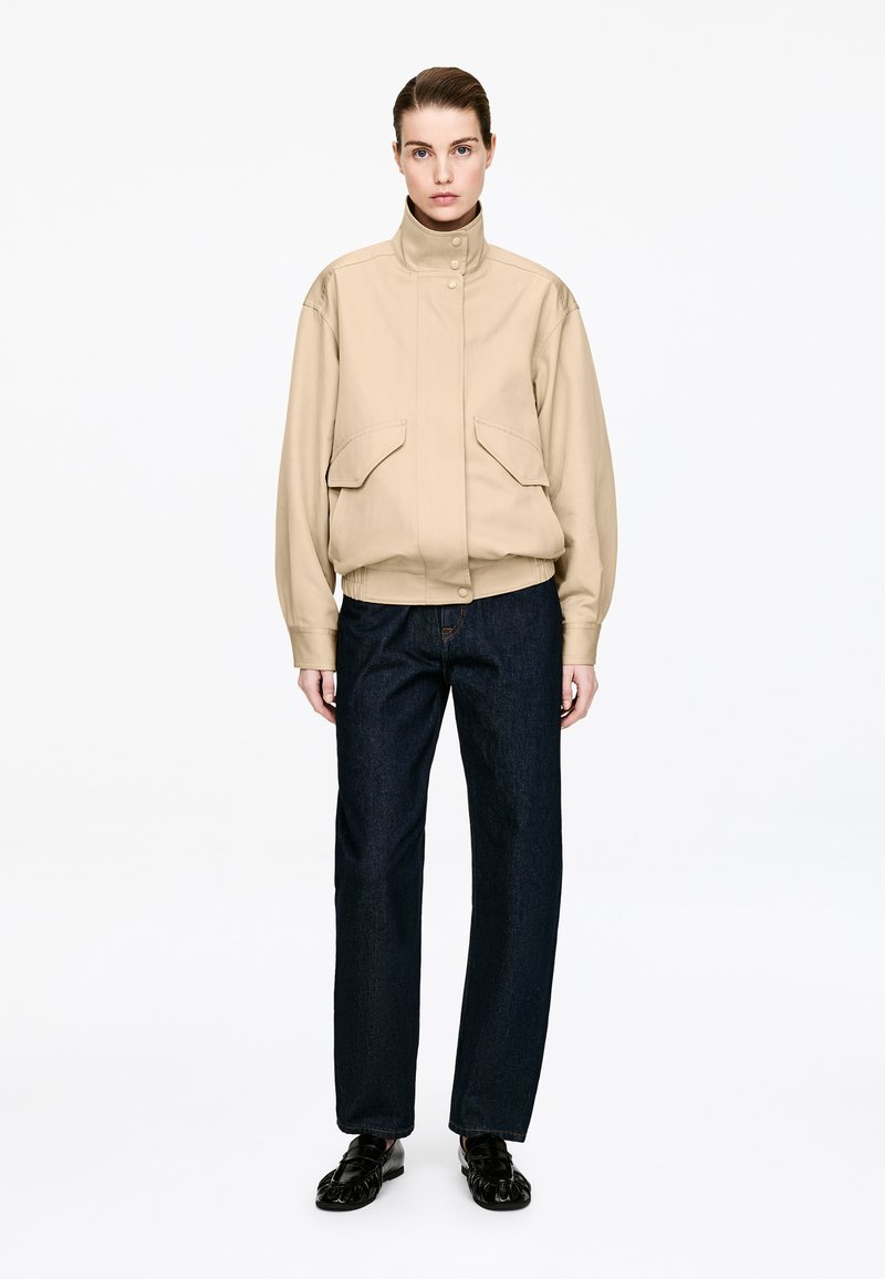 Femme portant une veste beige à col haut, un jean bleu foncé et des chaussures noires, debout face à l'avant sur un fond blanc.