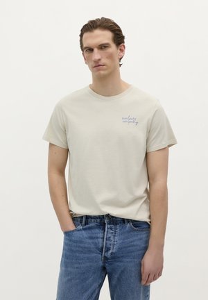 Beige tričko s krátkým rukávem, na kterém se na levé části hrudi nachází modrý text "scafers company", v kombinaci s modrými džínami.