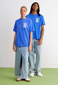 Zilas kokvilnas t-krekls ar baltu "OBEY World Wide Records" druku, valkāts ar gaismas denim svārkiem un biksēm. Baltas sporta kurpes papildina apģērbu.