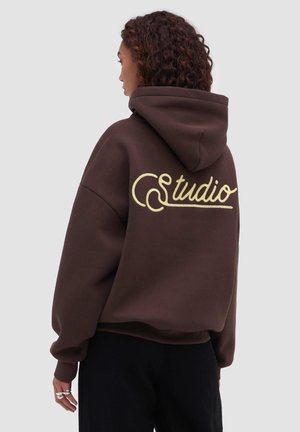 Kvinde med krøllet hår iført en brun hættetrøje med gul "Studio"-skrift på ryggen, stående mod en ensfarvet baggrund.