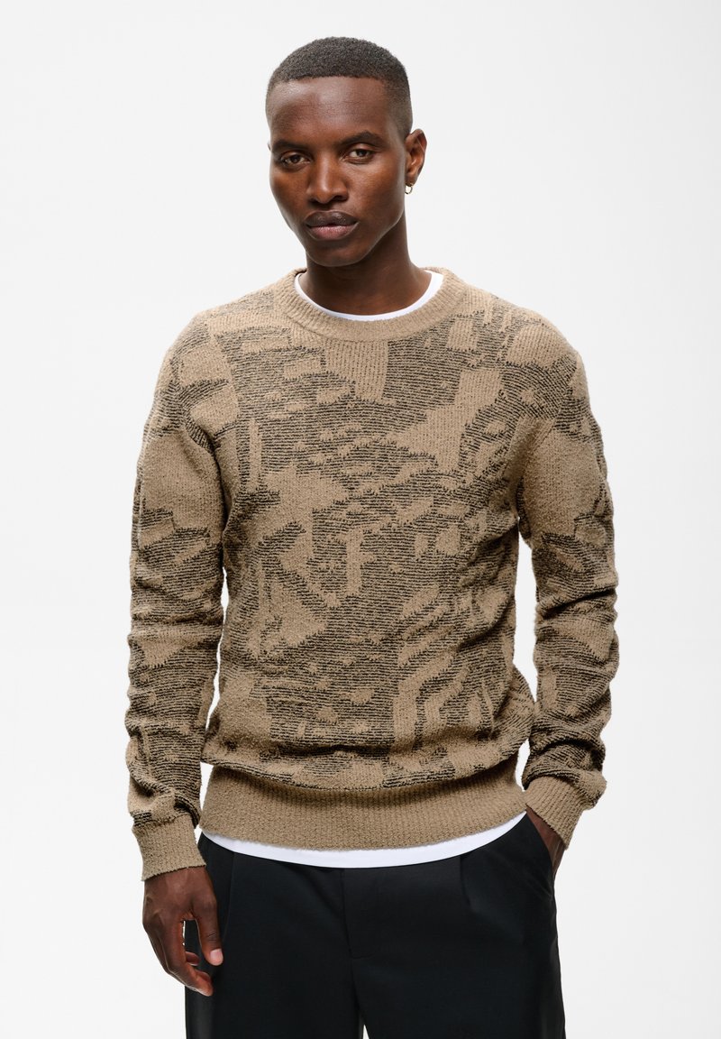 Homme portant un pull beige et marron à motifs par-dessus une chemise blanche avec un pantalon noir, debout devant un fond blanc uni.