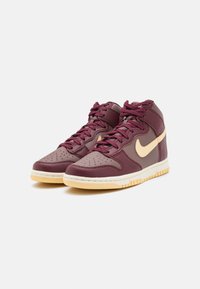 Tenisky s vysokým klínem z kaštanově červené kůže, béžové detaily a perforovaná špička. Mají silné tkaničky a světle hnědou gumovou podrážku.