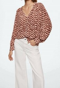 Donna che indossa una blusa lunga a maniche lunghe con motivo a chevron marrone e beige, abbinata a pantaloni bianchi a vita alta e gamba ampia.