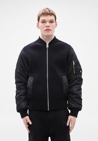 Casaco bomber preto com textura canelada no corpo e mangas em tecido liso. Apresenta um fecho de correr frontal, dois bolsos laterais e um bolso na manga.