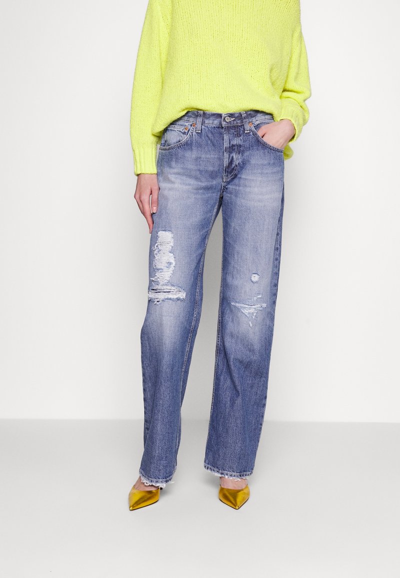 Personne portant un jean droit bleu clair déchiré, un pull jaune fluo et des chaussures pointues dorées métalliques, debout avec une main dans la poche.