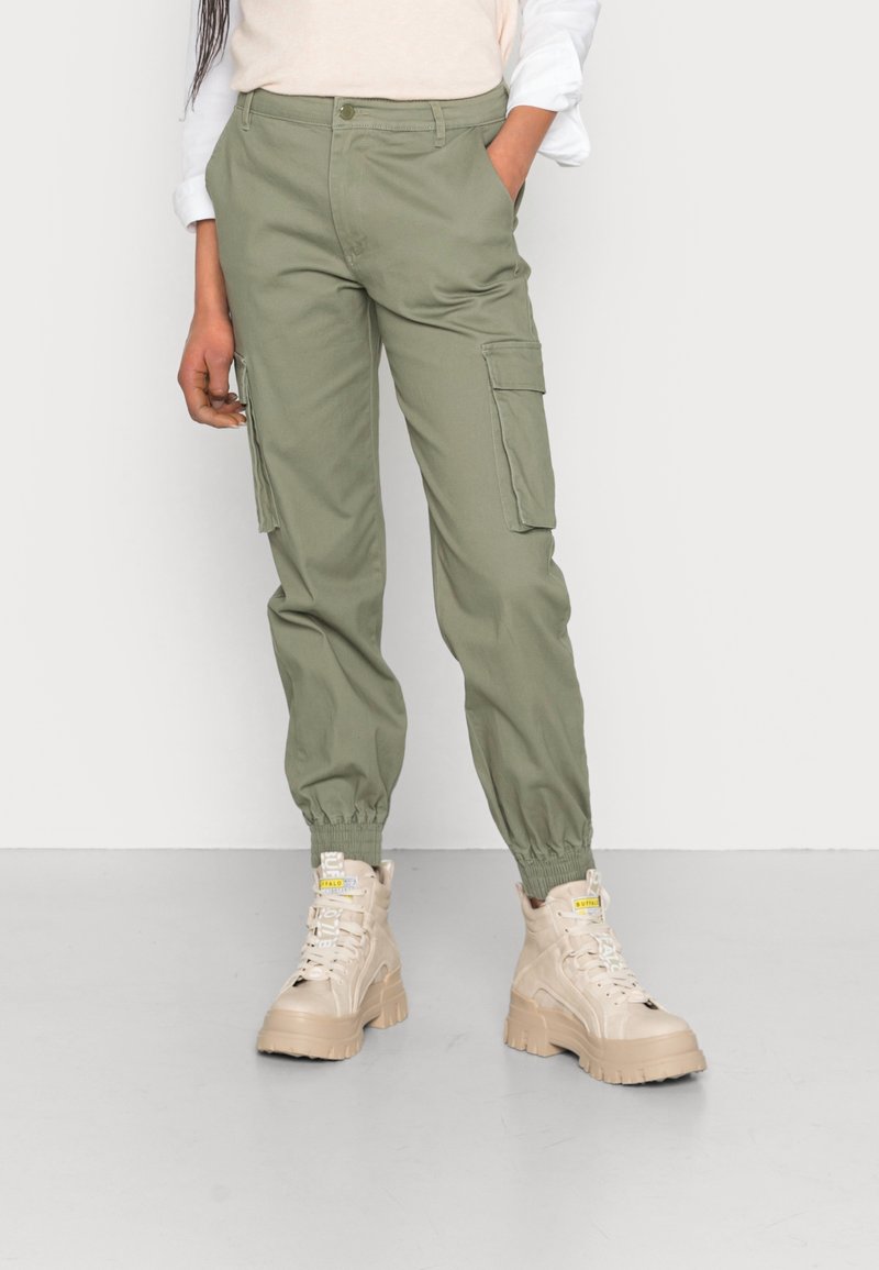 Personne portant un pantalon jogger cargo vert olive et des baskets montantes beige à semelle épaisse sur fond blanc.