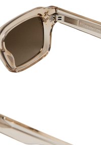 Ochelari de soare din acetat cu o ramă transparentă de culoare maro deschis, lentile maro închise și accente metalice pe balamale, având o formă modernă pătrată.