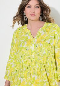Żółta koszula w kwiaty z rękawami do połowy przedramienia, zapinana na guziki, z marszczonym pasem. Ozdobiona żywym wzorem liści na lekkiej tkaninie.
