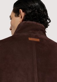 Brun mocka jacka med hög krage, med en läderetikett på baksidan och rena sömnadsdetaljer. Slät yta, minimalistisk design.