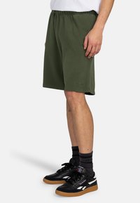 Green cotton shorts with an elastic waistband, featuring a relaxed fit and side pockets, paired with black sneakers and striped socks.