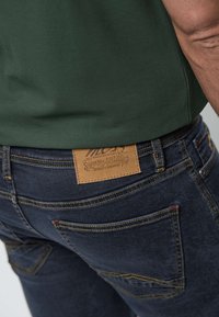 Rückansicht einer Person, die dunkelblaue Jeans mit braunem Lederetikett und ein grünes, eingestecktes Hemd trägt.