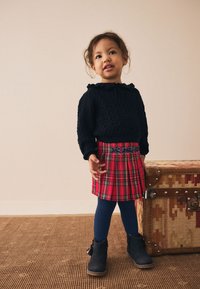 Marinblå texturerad tröja, röd plisserad kjol med rutmönster och blå accenter, blå stickade strumpbyxor och mörka stövlar. Placering mot en neutral bakgrund.