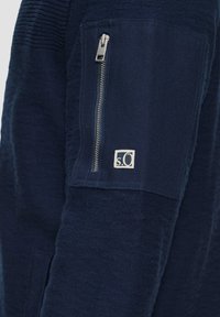 Marinefarbener Zip-Hoodie aus strukturiertem Stoff, mit einer Reißverschlusstasche an den Ärmeln und einem kleinen Logo im Taschenbereich.