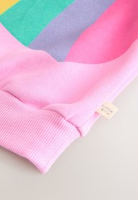 Roza jopa z večbarvnimi geometričnimi črtami v bleščeči zeleni, vijolični, rumeni in roza barvi; rebrasti manšeti in znamkasta etiketa.