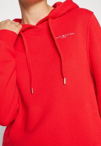 Sudadera de algodón roja con bolsillo frontal, capucha con cordones y extremos metálicos, y logo bordado en el pecho. Tejido texturizado.