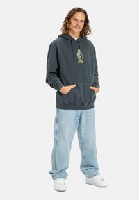 Tmavošedá mikina s kapucňou a prednými vreckami, s neónovým grafickým motívom. V kombinácii s voľnými svetlomodrými džínsami a bielymi teniskami.