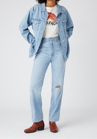Mujer que lleva una chaqueta de mezclilla azul claro y jeans con un desgarro en la rodilla derecha, camiseta blanca con diseño gráfico y botas de tobillo marrones.