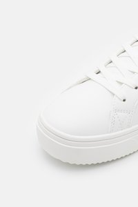Zapatilla blanca con una parte superior sintética suave, puntera redonda, cordones planos y una suela de goma texturizada con una plataforma ligeramente elevada.