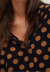 Tumme kankaita oleva paitapusero, jossa on suuria, pyöreitä, kultaruskeita pallokuvioita. Pehmeä, tekstuuriinen materiaali rentoa kaula-aukkosuunnittelua.