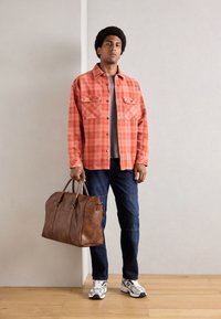Camisa de cuadros en rojo y naranja, camiseta gris debajo, jeans azules, bolsa de cuero marrón, zapatillas deportivas blancas y grises.