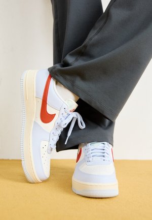 Białe i beżowe buty sportowe z czerwonym logo Nike i białymi sznurówkami, noszone z ciemnoszarymi spodniami i białymi skarpetkami na beżowej podłodze.
