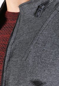 Chaqueta gris texturizada con un cuello alto y cremallera, que presenta un patrón sutil y una capa interior de punto roja visible en el escote.