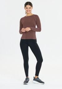 Ruskea pitkähihainen paita, jossa on raidalliset sivut, yhdistetty mustiin legginsseihin ja harmaisiin liikuntakenkiin. Malli seisoo tasaisella taustalla.