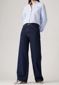 Chemise à boutons bleu clair, jean large bleu foncé, texture lisse, longueur totale, avec coutures visibles et un logo subtil en haut.