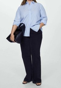 Chemise boutonnée à rayures bleu clair, pantalon évasé en denim foncé, tenant un sac noir. La chemise a une coupe décontractée et des manches retroussées.