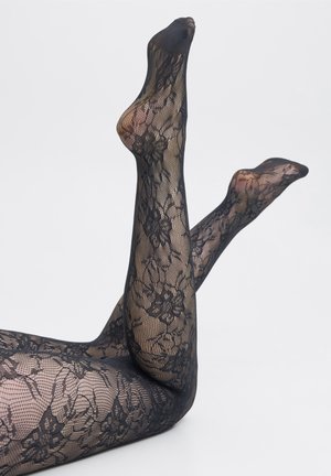 Jambes levées en collants noirs à motif de dentelle florale sur fond blanc.